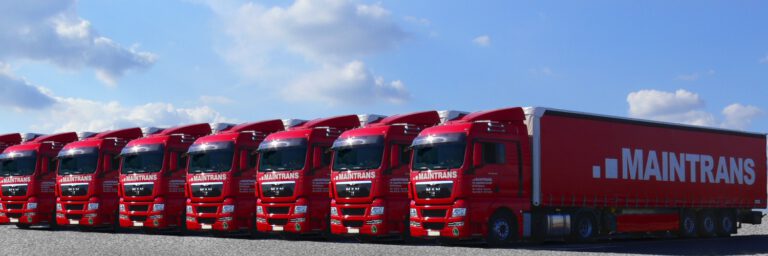 Acht Maintrans-LKW stehen nebeneinander auf einem Parkplatz, im Hintergrund blauer Himmel und Sonne.