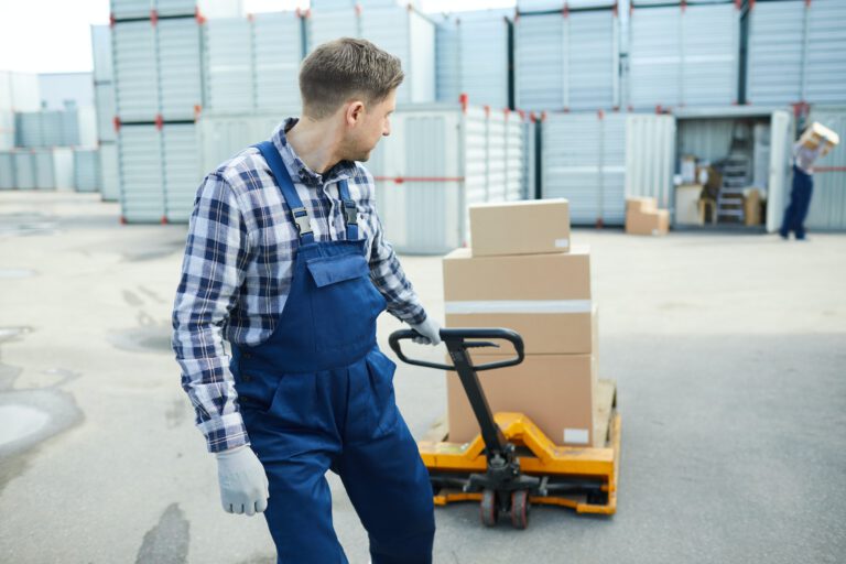 Ein Lagermitarbeiter in blauer Latzhose zieht einen Hubwagen mit Kartons über den Hof eines Logistikunternehmens.