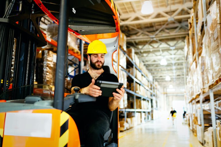 Ein Lagermitarbeiter sitzt auf seinem Gabelstapler im Logistik-Lager und sieht sich seinen Kommissionierungs-Auftrag auf dem Tablet an.