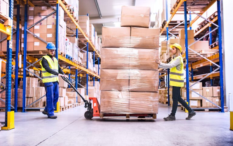 Ein Lagermitarbeiter und eine Lagermitarbeiterin ziehen einen Hubwagen mit verpackter Ware auf Palette durch ein Logistik-Lager.