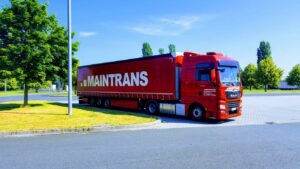 Ein roter Maintrans-LKW mit weißem Maintrans-Logo steht auf einem Parkplatz neben einem grünen Rasenstück mit Bäumen.