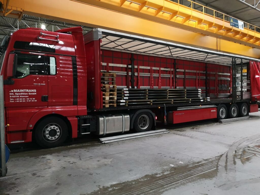 Ein Maintrans-LKW mit seitlich geöffneter Auflieger-Plane hat Langgut als Teilladung geladen.