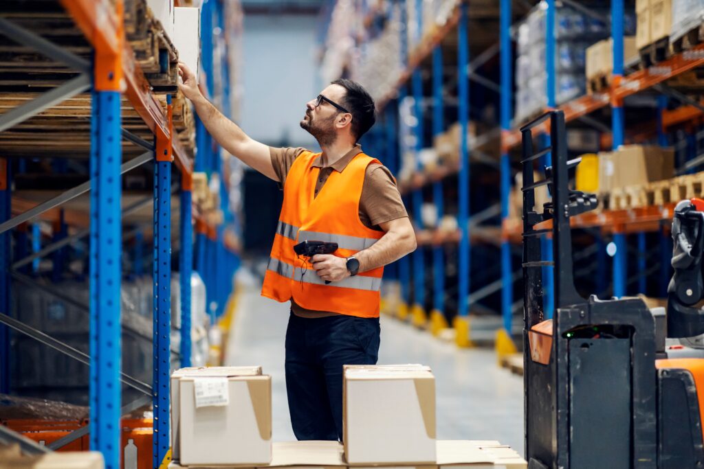 Ein Lagermitarbeiter steht vor einem Hochregal ind einem Logistik-Lager und scannt eingelagerte Waren mit dem Handscanner ein.