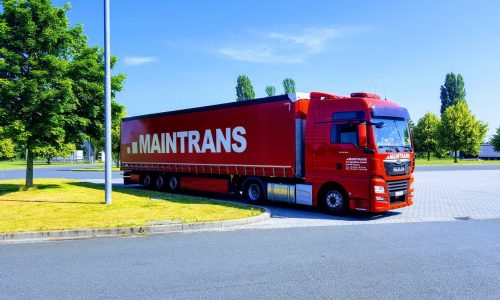 Ein roter Maintrans-LKW mit weißem Maintrans-Logo steht auf einem Parkplatz neben einem grünen Rasenstück mit Bäumen.