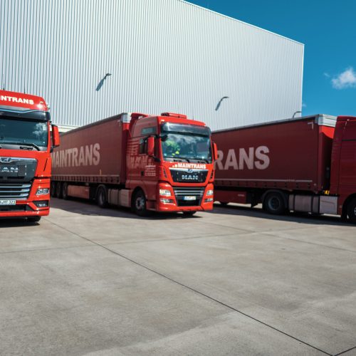 Drei rote LKW mit Maintrans Logo stehen nebeneinander an der Laderampe eines Logistik-Lagers.